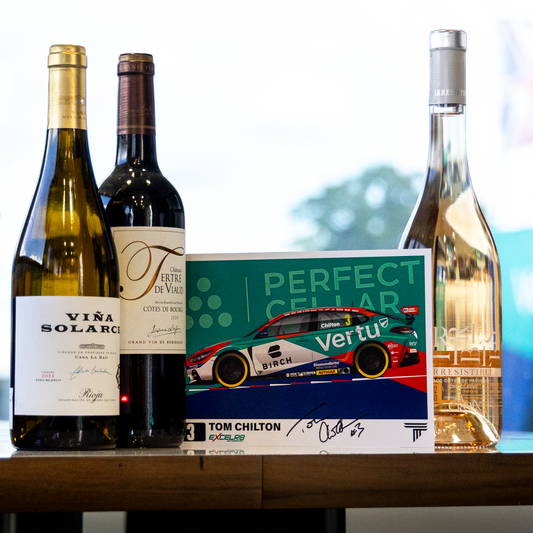 Three wine bottles and a book titled 'Perfect Cellar' with Tom Chilton's Vertue edition, arranged on a wooden table in front of a bright window.