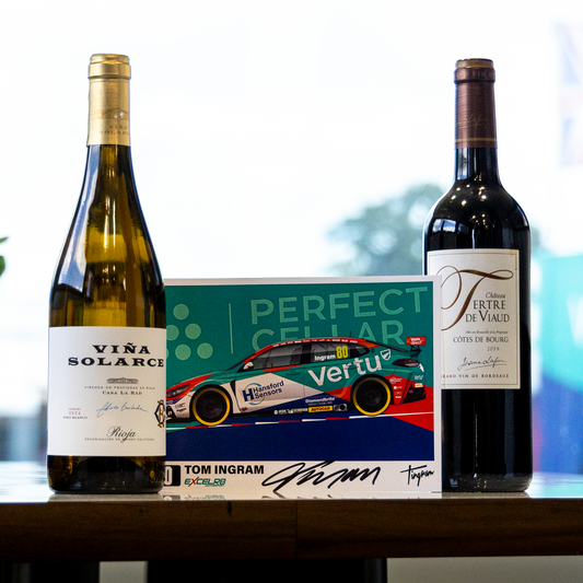 Two wine bottles and a branded box displayed on a table in front of a bright window. The left bottle is a pale green glass bottle labeled VIVA SOLARIUS, the right bottle is a dark red wine bottle labeled VINTI E VINO, and the center is a box with a race car graphic and the text PERFECT CELAR VERTU TOM INGRAM.