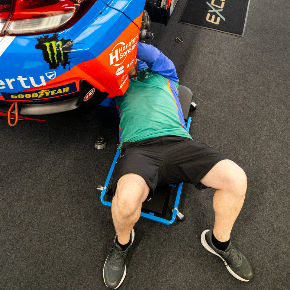 A mechanic in a green shirt and black shorts lies on the Car Creeper & Seat by Draper tools