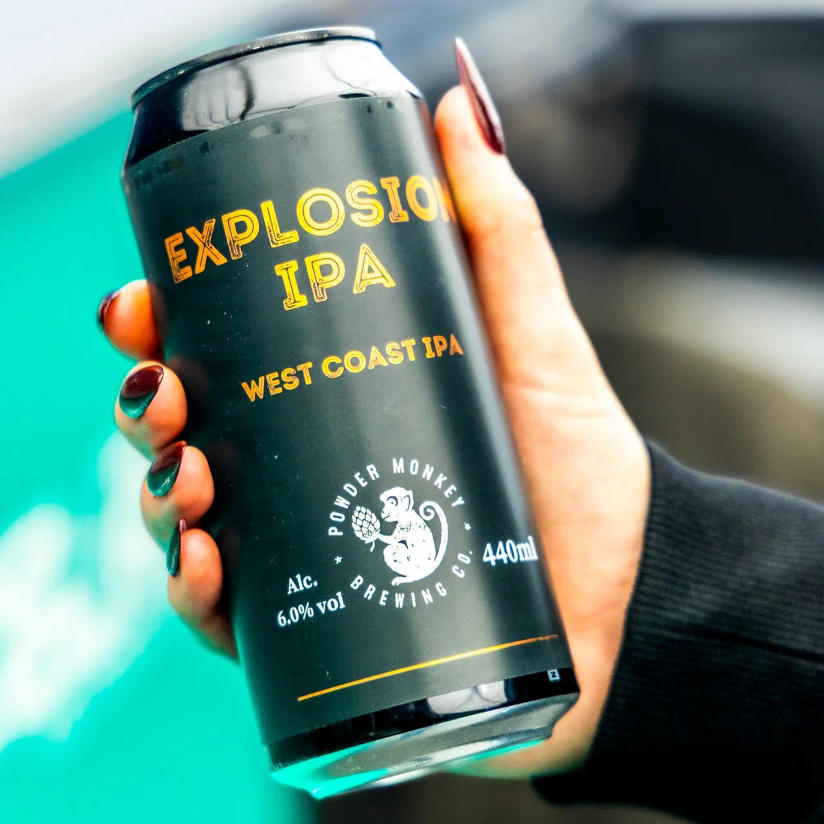A hand with dark red nail polish holds a black can of Explosion IPA West Coast IPA 