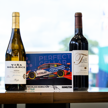 Two wine bottles and a signed card on a wooden table. The left bottle is a pale golden white wine labeled VIRA SOLARCE, the right bottle is a dark red wine, and the center is a blue and red Perfect Cellar signed card featuring racer Nicholas Hamilton.