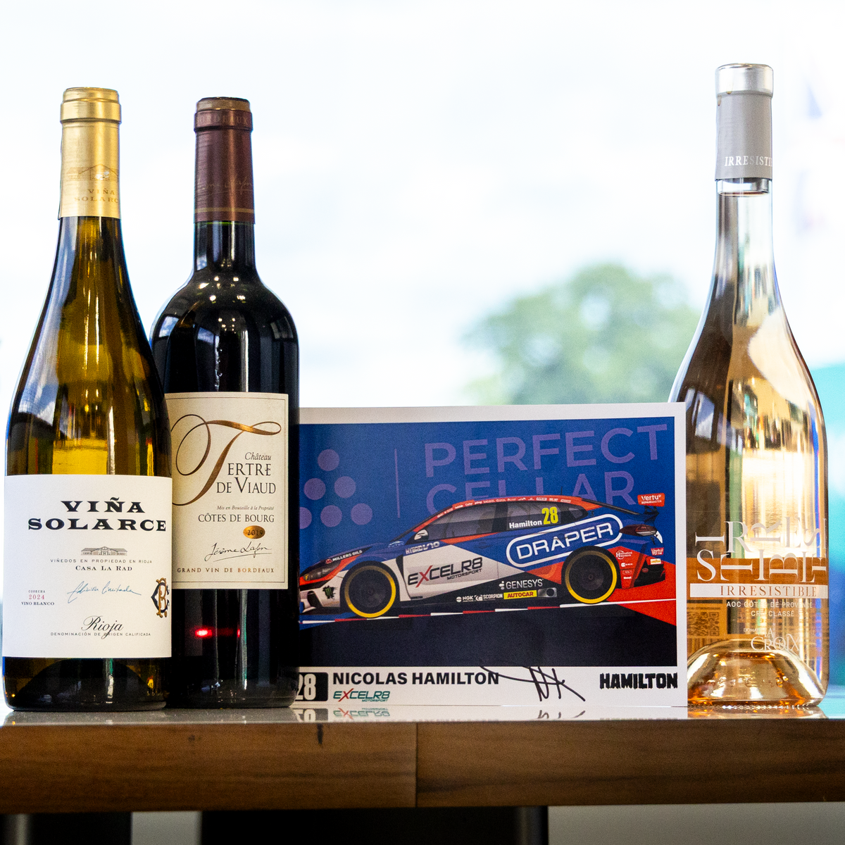 Three wine bottles and a branded box arranged on a wooden table indoors. Visible products include a white wine bottle labeled Vira Solarese, a dark red wine bottle with a large 'F' label, a clear rosé wine bottle with a silver cap, and a box featuring a race car and the text 'Perfect Gear' and 'Nicolas Hamilton'.