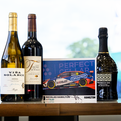 A collection of premium bottles and a collectible card displayed on a wooden table. The collection includes a bottle of Vira Solarece white wine, a bottle of Ferito di Vico red wine, a Perfect Cellar Nicolas Hamilton card, and a bottle of Perseus 13% ABV sparkling wine with a black patterned label.
