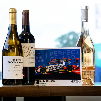 Three wine bottles and a printed card or box standing on a wooden table in a real indoor setting with a window and plant visible in the background. The bottles appear to be the product being shown, with one white wine bottle labeled VIVA SOLARCE, one dark red wine bottle with a large script logo, and one pale pink wine bottle with a metallic cap.