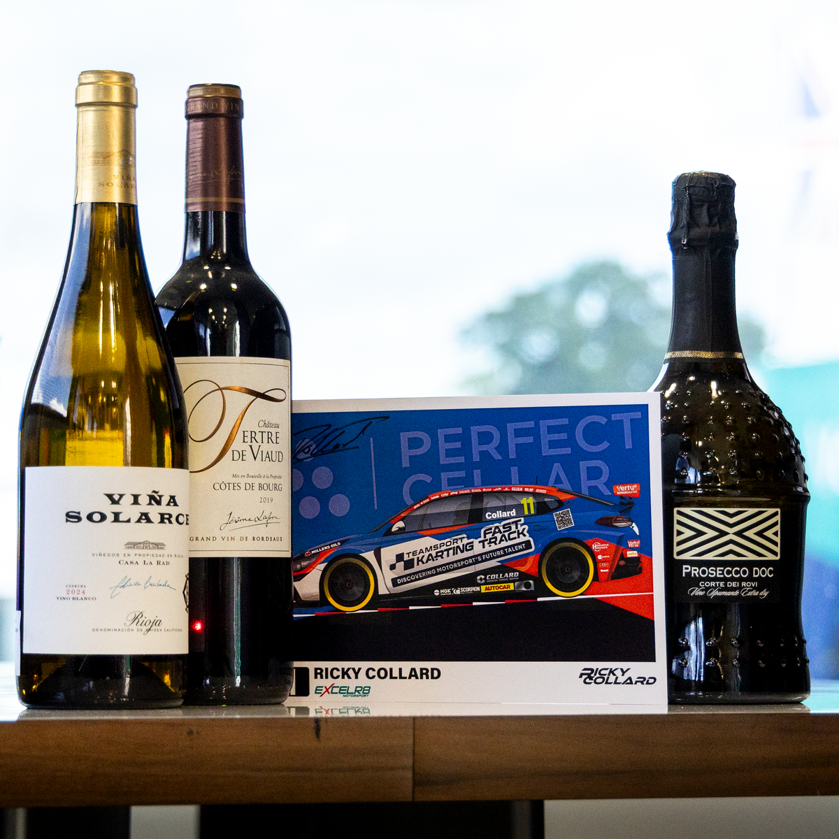 A collection of gourmet beverages arranged on a wooden table, featuring a bottle of Vira Solari white wine, a bottle of Valtori wine, a Proton Drive sparkling wine bottle, and a race card for Ricky Collard.
