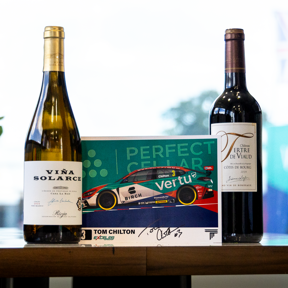 Two wine bottles and a signed collectible card displayed on a wooden table in a bright indoor setting. The left bottle is a white wine bottle labeled 'VISA SOLARIS', the right bottle is a dark red wine bottle labeled 'Ferre d'Avion', and the center is a green and red 'Perfect Center' signed card featuring Tom Chilton signatures.