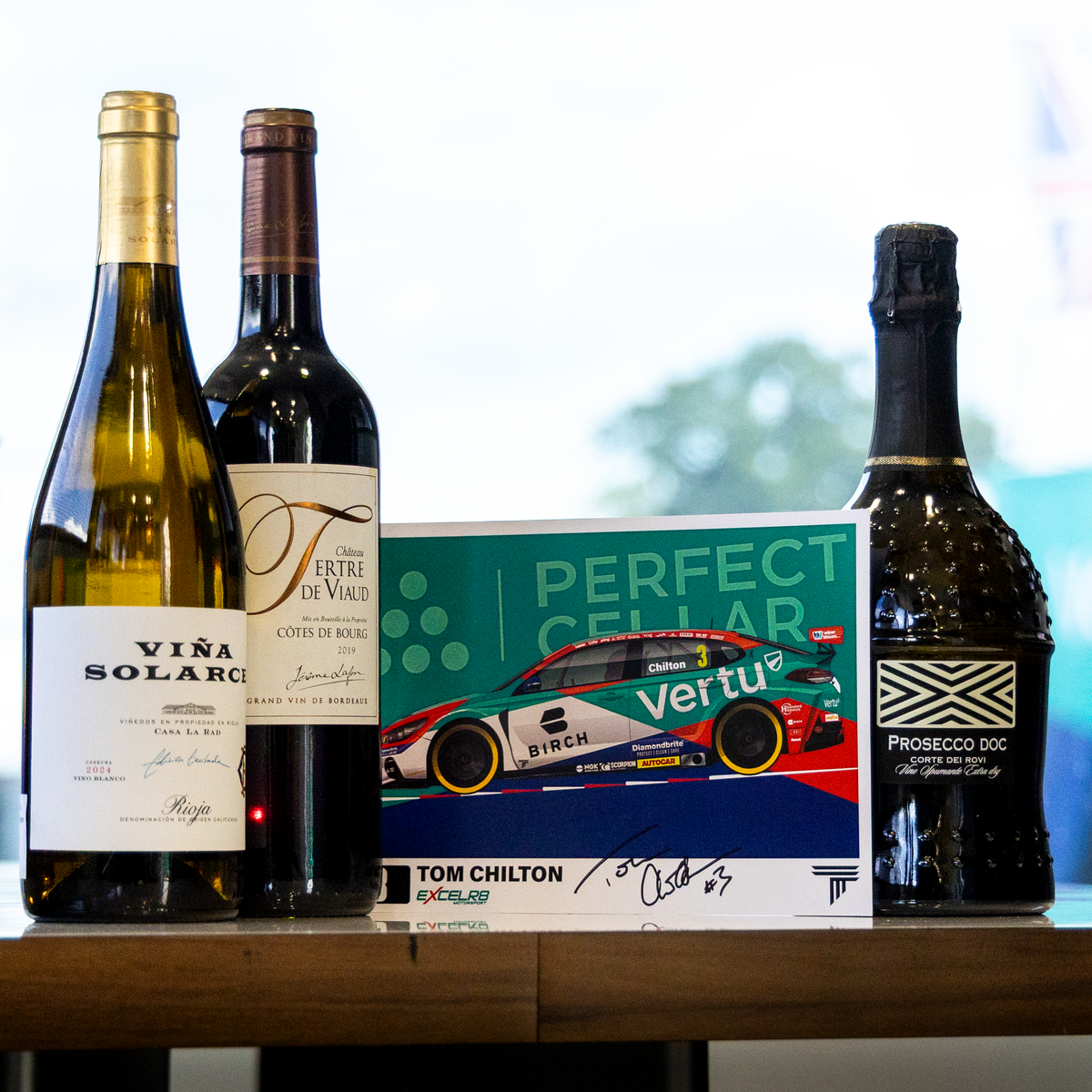 A curated selection of gourmet bottles and a signed collectible card displayed on a wooden table in a modern room setting. The collection includes a bottle of Vira Solarce white wine, a bottle of Perugino di Valtellino wine, a bottle of Vertu race car champagne, and a signed Tom Chilton card.