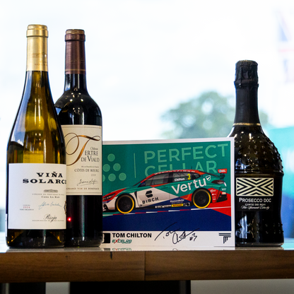 A curated selection of gourmet bottles and a signed collectible card displayed on a wooden table in a modern room setting. The collection includes a bottle of Vira Solarce white wine, a bottle of Perugino di Valtellino wine, a bottle of Vertu race car champagne, and a signed Tom Chilton card.