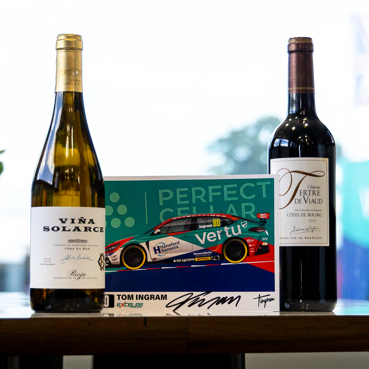 Two wine bottles and a branded box displayed on a table in front of a bright window. The left bottle is a pale green glass bottle labeled VIVA SOLARIUS, the right bottle is a dark red wine bottle labeled VINTI E VINO, and the center is a box with a race car graphic and the text PERFECT CELAR VERTU TOM INGRAM.