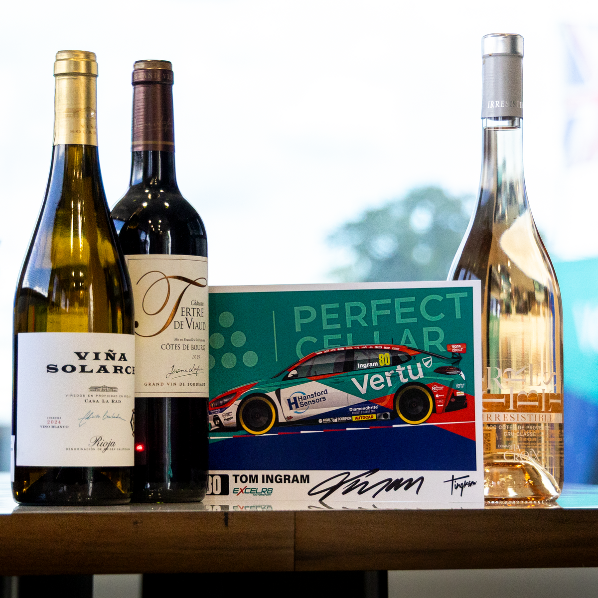 A curated selection of wine bottles and a signed collectible card on a wooden table. The collection includes a green bottle of Vira Solari white wine, a dark red bottle of Conifera Riserva red wine, a signed Tom Ingdran 'Perfect Cellar' collectible card, and a bottle of rosé wine, set against a softly blurred indoor background.