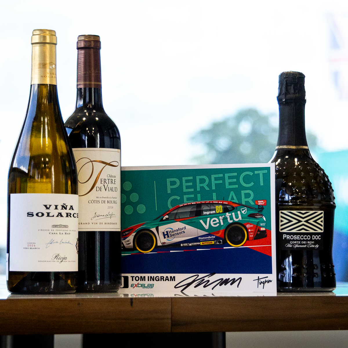 A curated collection of premium bottles and a signed collectible card displayed on a wooden table. The set includes a bottle of Vira Solari white wine, a bottle of Valtori red wine, a bottle of Prosecco, and a signed 'Perfect Cellar' Tom Ingram card.